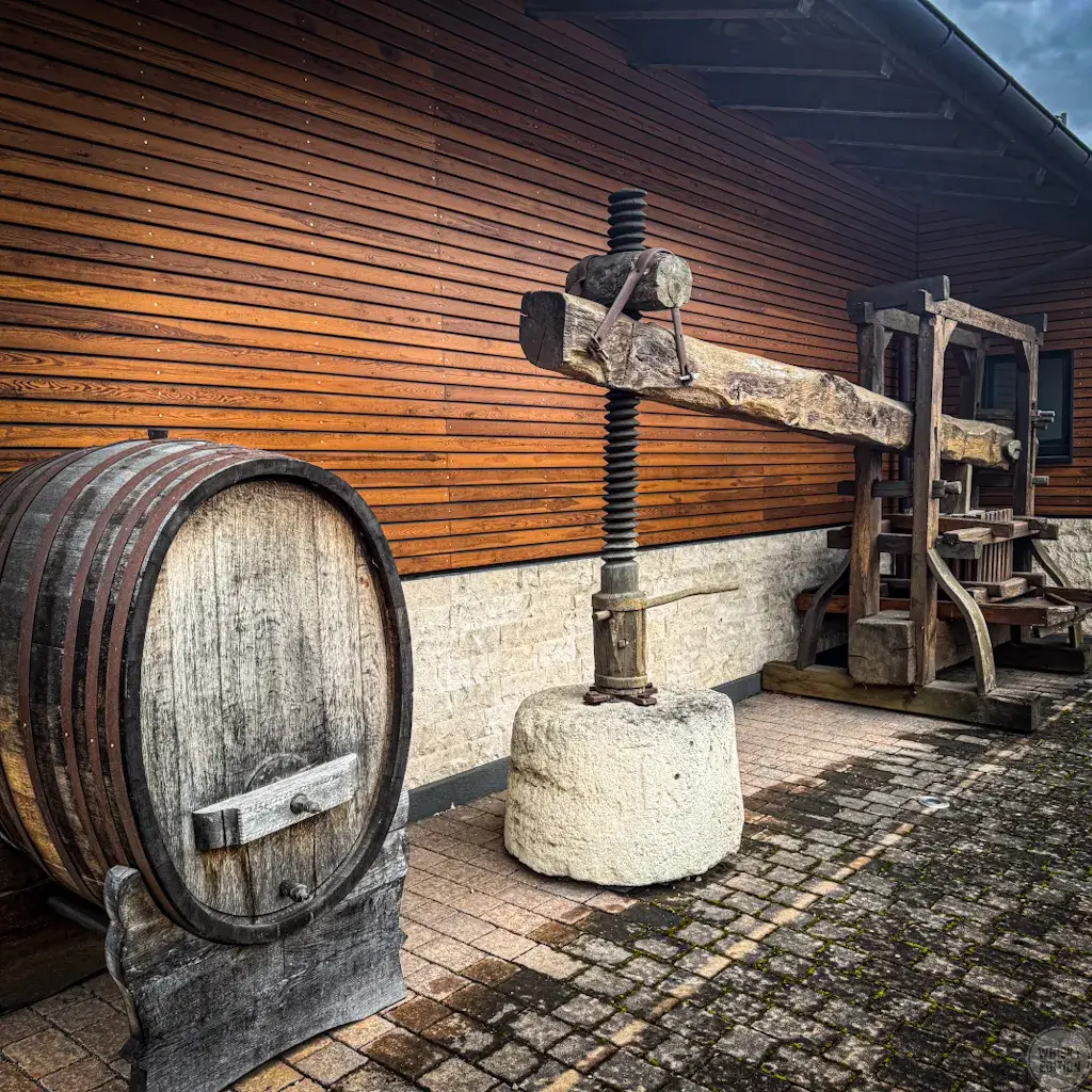 Historische Weinpresse und altes Fass vor dem Holzgebäude der Destillerie