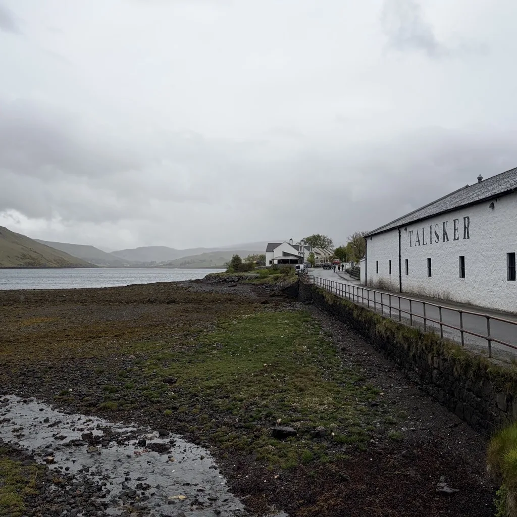 Die Talisker Distillery am Ufer des Loch Harport bei grauem Wetter, im Hintergrund die Hügel von Skye